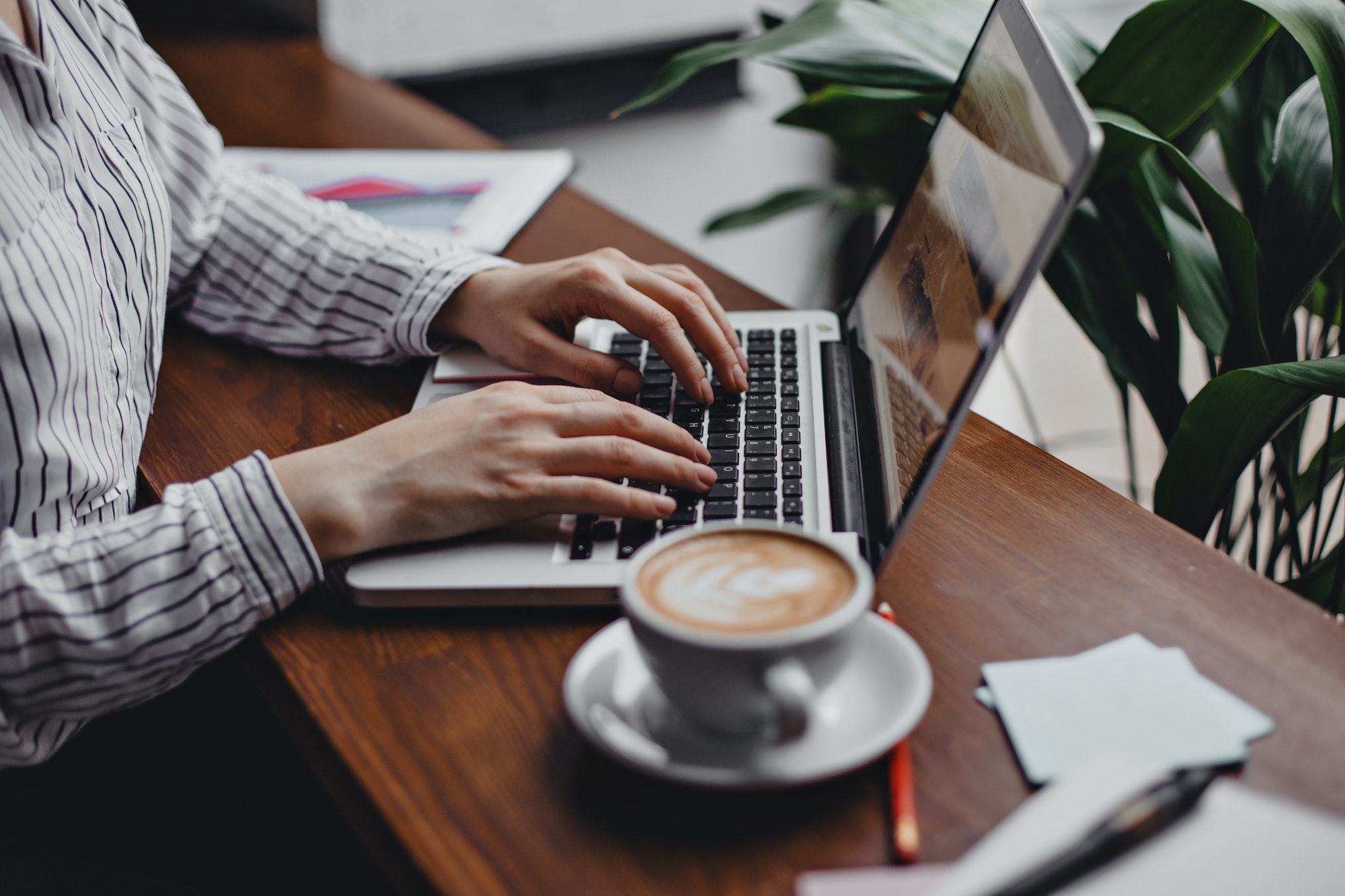 Woman works in laptop while sitting in cozy office at table. Shot of cup of coffee, sheets of paper